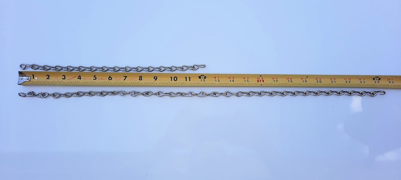 May include: Two silver chains, one measuring 25 cm and the other measuring 63.5 cm, are laid out on a white surface next to a yellow measuring tape.