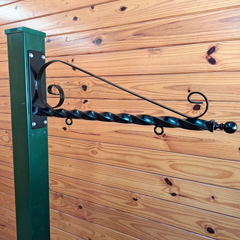 Op de afbeelding: Een zwarte smeedijzeren bordenhouder met een gedraaid staafontwerp. De bordenhouder is gemonteerd op een donkergroene paal en heeft decoratief krulwerk en een kogelvormige bekroning. De achtergrond is een houten muur.