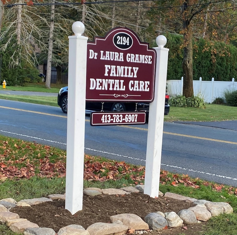 Puede incluir: Un letrero rectangular granate con texto blanco que dice 2194, Dr LAURA GRAMSE, FAMILY DENTAL CARE y 413-783-6907. El letrero est&aacute; montado entre dos postes blancos rematados con esferas, en un parterre bordeado por piedras grises.