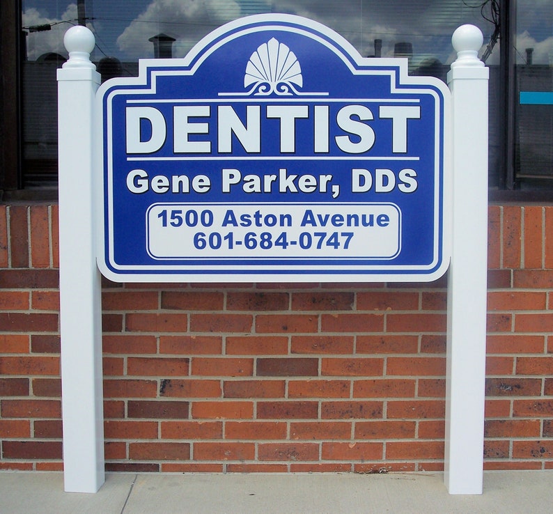 Puede incluir: Un letrero rectangular azul y blanco con una parte superior arqueada decorativa y un icono de concha. El letrero dice DENTIST, Gene Parker, DDS, 1500 Aston Avenue y 601-684-0747. Est&aacute; montado entre dos postes blancos con remates esf&eacute;ricos.