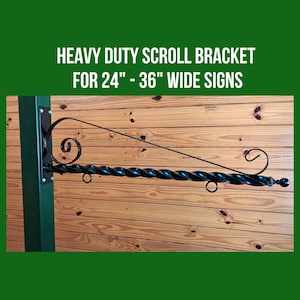 May include: A black, heavy-duty scroll bracket for signs 24"-36" wide. The bracket has a twisted bar design with decorative scrollwork and a ball finial. The text "HEAVY DUTY SCROLL BRACKET FOR 24" - 36" WIDE SIGNS" is displayed.
