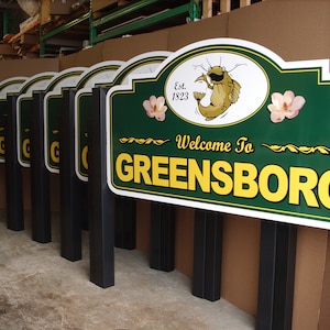 Puede incluir: Una fila de letreros rectangulares verdes con bordes blancos y postes negros. Cada letrero presenta el texto Welcome To GREENSBORO en amarillo, una ilustraci&oacute;n de un bagre, el texto Est. 1823 y dos flores rosas como acentos.