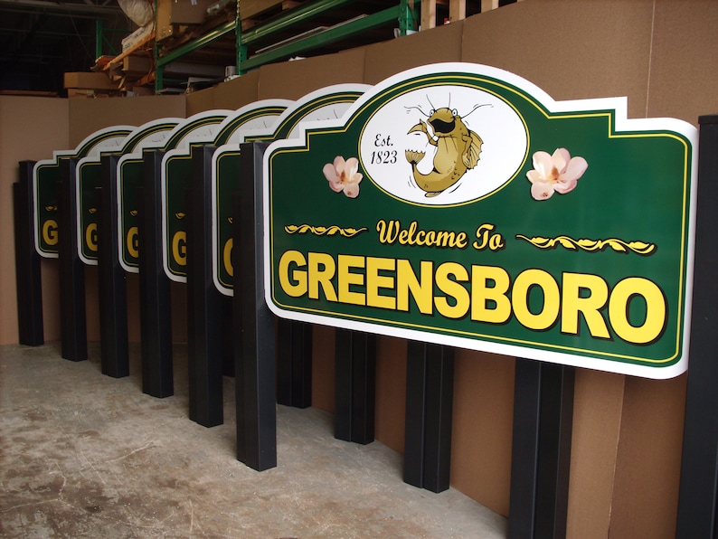 Puede incluir: Una fila de letreros rectangulares verdes con bordes blancos y postes negros. Cada letrero presenta el texto Welcome To GREENSBORO en amarillo, una ilustraci&oacute;n de un bagre, el texto Est. 1823 y dos flores rosas como acentos.