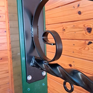 Op de afbeelding: Zwarte smeedijzeren beugel met een decoratief krulontwerp, gemonteerd op een groene paal met een zwarte metalen plaat. De beugel is bevestigd met zilveren bouten en is geschikt voor het ophangen van items.