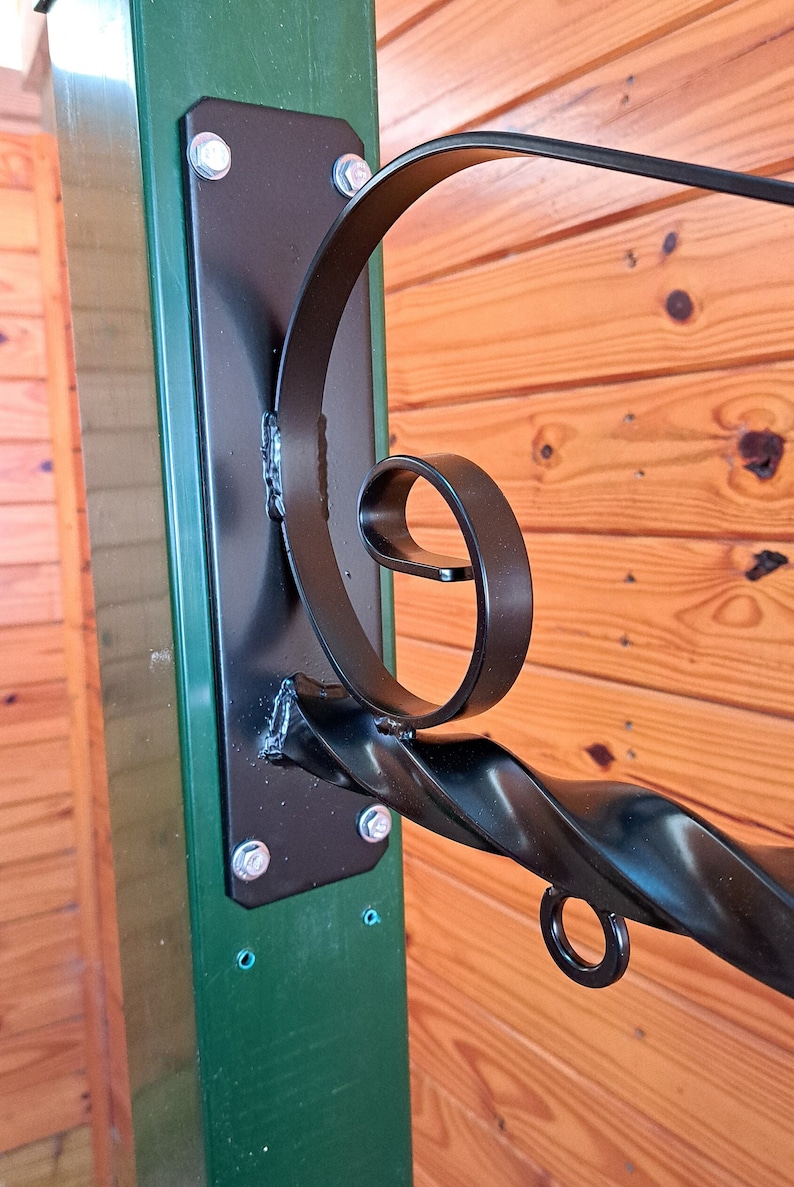 Op de afbeelding: Zwarte smeedijzeren beugel met een decoratief krulontwerp, gemonteerd op een groene paal met een zwarte metalen plaat. De beugel is bevestigd met zilveren bouten en is geschikt voor het ophangen van items.
