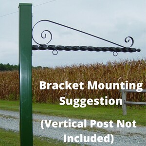 Op de afbeelding: Een donkergroene metalen beugel met een decoratieve gedraaide staaf en krulontwerp. De beugel is gemonteerd op een groene verticale paal. De tekst op de afbeelding luidt "Bracket Mounting Suggestion (Vertical Post Not Included)".