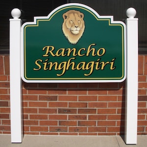 Puede incluir: Un letrero rectangular verde con borde blanco y adornos dorados, montado sobre dos postes blancos con remates esf&eacute;ricos. El letrero presenta un gr&aacute;fico de una cabeza de le&oacute;n en la parte superior y el texto "Rancho Singhagiri" en letras doradas.