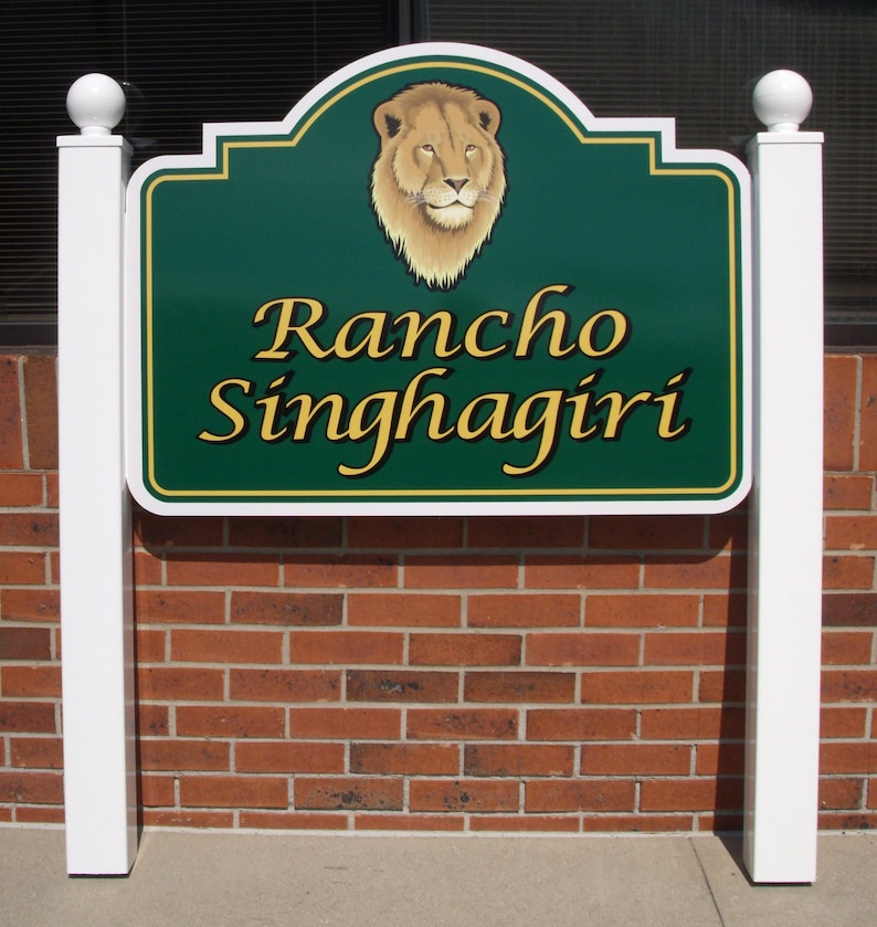 Puede incluir: Un letrero rectangular verde con borde blanco y adornos dorados, montado sobre dos postes blancos con remates esf&eacute;ricos. El letrero presenta un gr&aacute;fico de una cabeza de le&oacute;n en la parte superior y el texto "Rancho Singhagiri" en letras doradas.