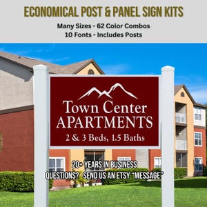 May include: A post and panel sign kit featuring a dark red rectangular sign with white text reading Town Center APARTMENTS 2 & 3 Beds, 1.5 Baths and a mountain graphic. The sign is mounted between two white vertical posts on a grassy lawn.