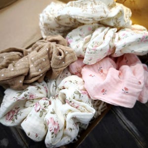 May include: A selection of fabric scrunchies in various colours and patterns. The scrunchies feature floral prints in pink and white, a brown scrunchie with white polka dots, and a cream-coloured scrunchie with a floral design. The scrunchies are in a cardboard box.