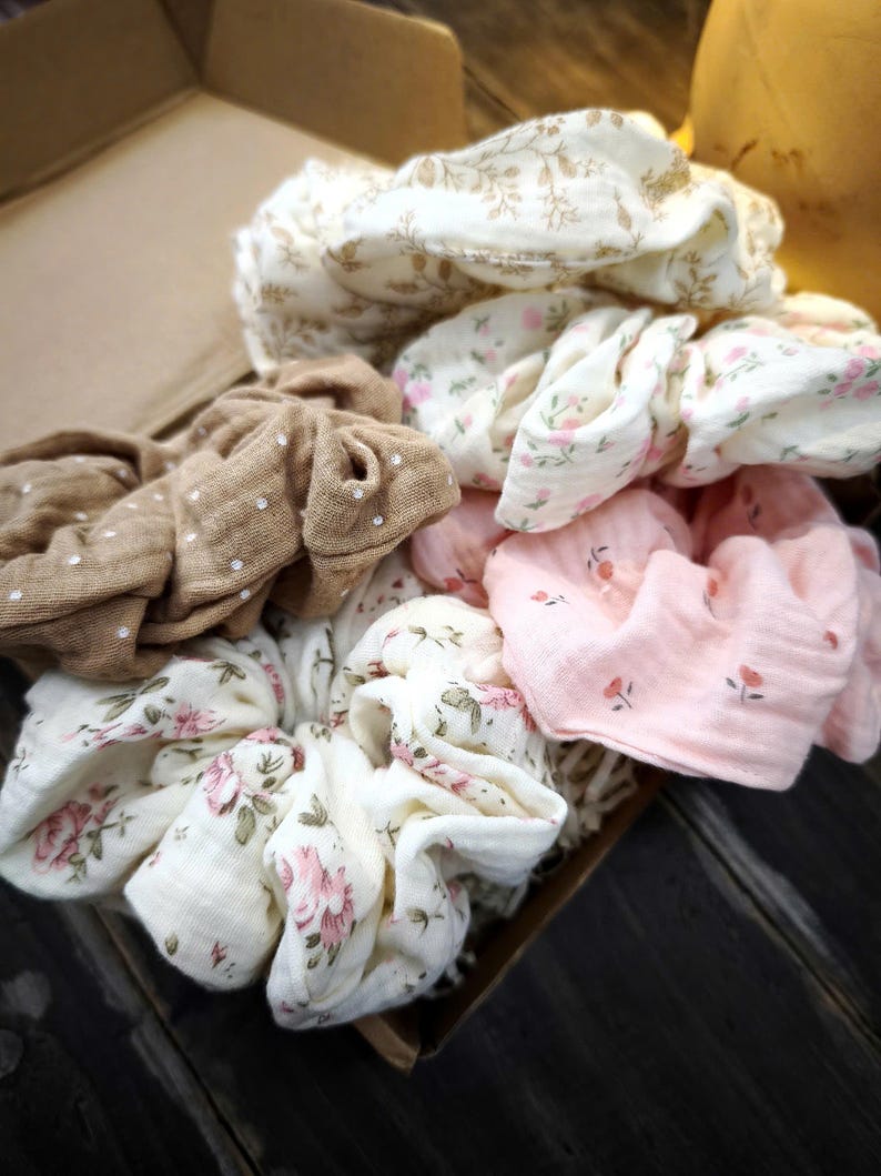May include: A selection of fabric scrunchies in various colours and patterns. The scrunchies feature floral prints in pink and white, a brown scrunchie with white polka dots, and a cream-coloured scrunchie with a floral design. The scrunchies are in a cardboard box.