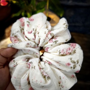 May include: A white scrunchie featuring a floral design of pink roses and green leaves. The fabric is gathered and bunched, creating a textured look. This hair accessory is designed to hold hair and adds a touch of feminine style.