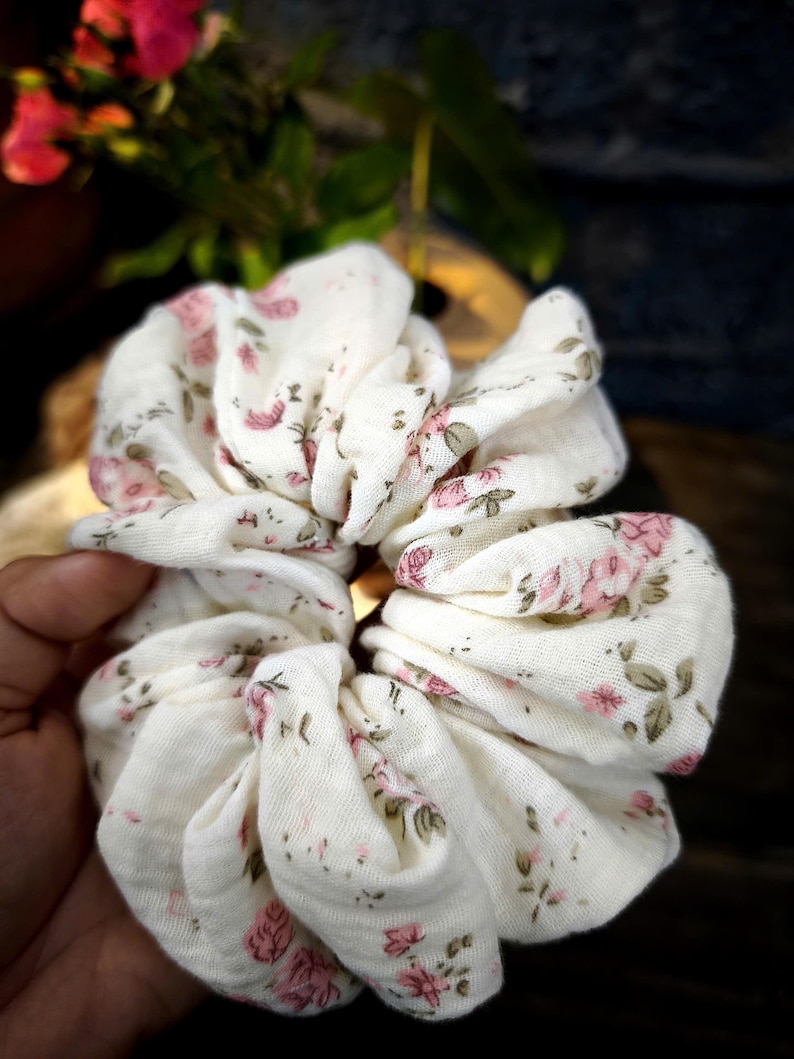 May include: A white scrunchie featuring a floral design of pink roses and green leaves. The fabric is gathered and bunched, creating a textured look. This hair accessory is designed to hold hair and adds a touch of feminine style.