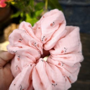 May include: A pale pink scrunchie with a floral design. The fabric is gathered, creating a full appearance. The small flowers are a muted red with dark green stems, adding a touch of charm to this hair accessory.