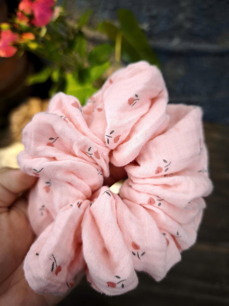 May include: A pale pink scrunchie with a floral design. The fabric is gathered, creating a full appearance. The small flowers are a muted red with dark green stems, adding a touch of charm to this hair accessory.