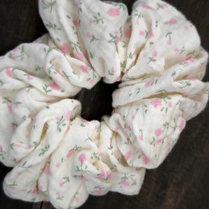 May include: A white scrunchie featuring a pattern of small pink roses and green leaves. The fabric is gathered, creating a voluminous hair accessory. The scrunchie is a hair accessory, perfect for adding a touch of floral charm.