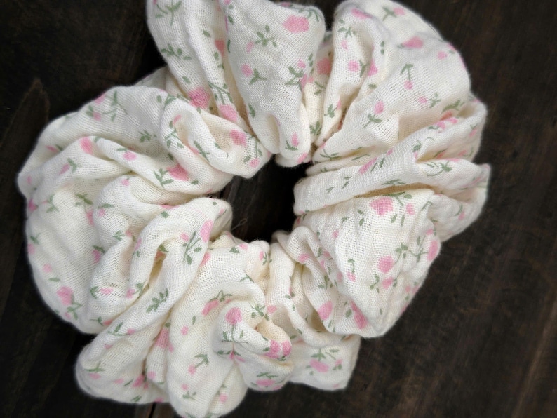 May include: A white scrunchie featuring a pattern of small pink roses and green leaves. The fabric is gathered, creating a voluminous hair accessory. The scrunchie is a hair accessory, perfect for adding a touch of floral charm.