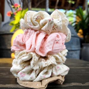 May include: A stack of fabric scrunchies in various colours and patterns. The scrunchies include floral prints in cream and pink, and solid pink. The base is a tan fabric with white polka dots.