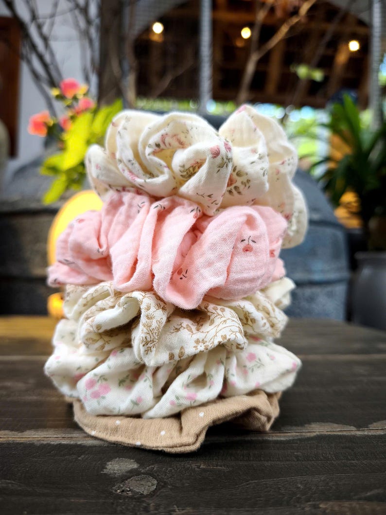 May include: A stack of fabric scrunchies in various colours and patterns. The scrunchies include floral prints in cream and pink, and solid pink. The base is a tan fabric with white polka dots.