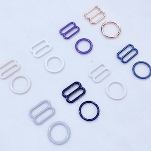 Op de afbeelding: Assortiment van metalen bh-schuifjes in verschillende kleuren, waaronder wit, paars, blauw en goud. De schuifjes zijn gerangschikt op een wit oppervlak.