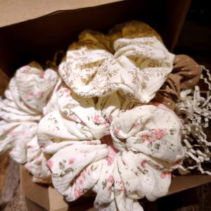 May include: A selection of fabric scrunchies presented in a cardboard box. The scrunchies display floral patterns in cream, pink, and brown. Some have a cream base with pink flowers, while others have a brown base with a floral design.