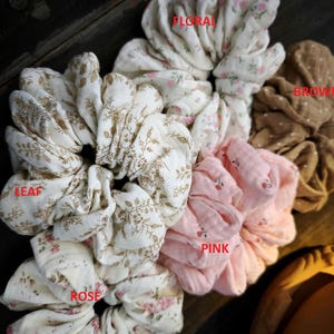 May include: A selection of fabric scrunchies in different colours and patterns. The scrunchies feature floral, leaf, rose, and polka dot designs. Colours include white, pink, and brown. The scrunchies are made of soft fabric.