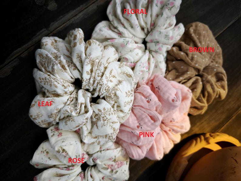 May include: A selection of fabric scrunchies in different colours and patterns. The scrunchies feature floral, leaf, rose, and polka dot designs. Colours include white, pink, and brown. The scrunchies are made of soft fabric.