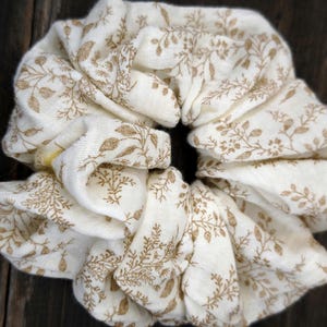 May include: An ivory scrunchie featuring a repeating pattern of brown floral sprigs. The fabric is gathered and folded, creating a voluminous, textured appearance. The scrunchie is designed for hair styling.
