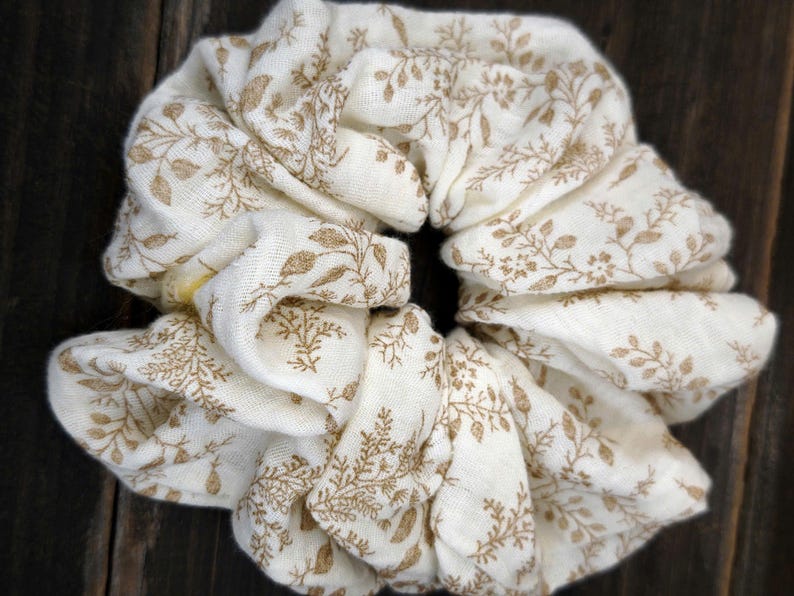 May include: An ivory scrunchie featuring a repeating pattern of brown floral sprigs. The fabric is gathered and folded, creating a voluminous, textured appearance. The scrunchie is designed for hair styling.