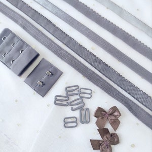Op de afbeelding: Assortiment van grijze elastische bh-bandjes, bh-haken en ogen en bruine satijnen strikken. De items zijn gerangschikt op een witte achtergrond met gouden glitter.