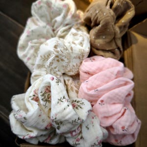 May include: A selection of fabric scrunchies in various colours and patterns. The scrunchies feature floral prints in pink and white, a brown scrunchie with white polka dots, and a plain pink scrunchie. The scrunchies are in a brown box.