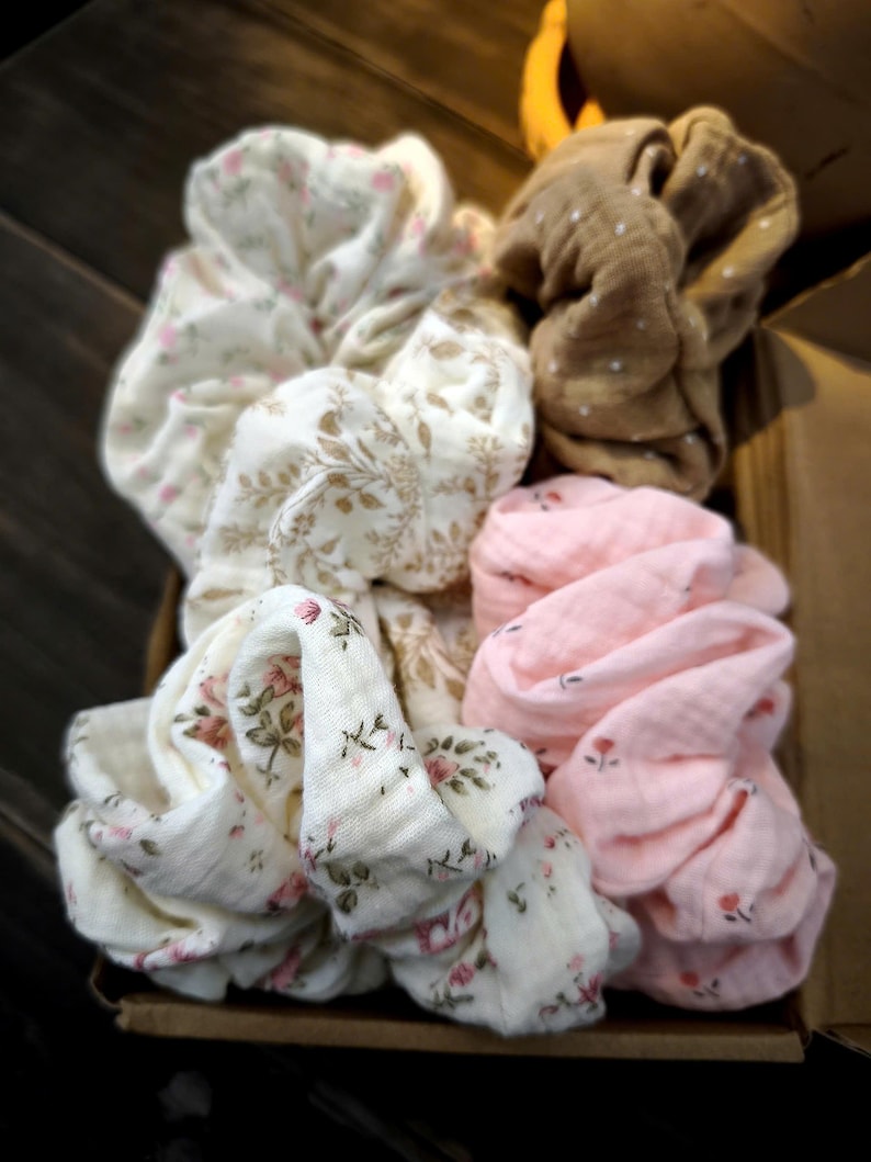 May include: A selection of fabric scrunchies in various colours and patterns. The scrunchies feature floral prints in pink and white, a brown scrunchie with white polka dots, and a plain pink scrunchie. The scrunchies are in a brown box.