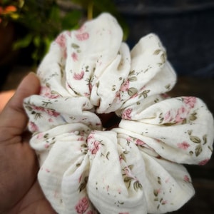 May include: A white scrunchie featuring a pattern of pink roses and green foliage. The fabric is gathered, creating a full, textured look. This hair accessory is designed to secure hair in a ponytail or bun, adding a touch of floral elegance.