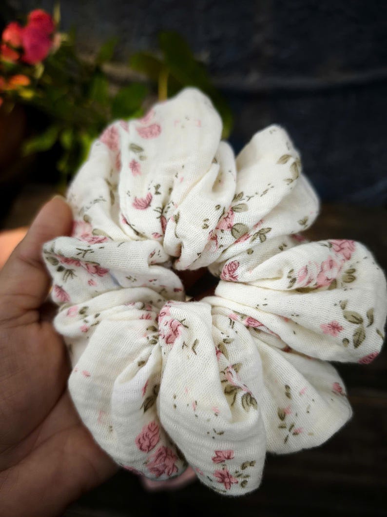 May include: A white scrunchie featuring a pattern of pink roses and green foliage. The fabric is gathered, creating a full, textured look. This hair accessory is designed to secure hair in a ponytail or bun, adding a touch of floral elegance.