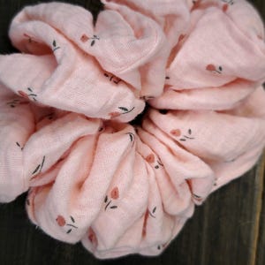 May include: A large, pale pink scrunchie with a floral design. The scrunchie is made of soft material and has small, dark green leaves and tiny red flowers. The scrunchie is designed to hold hair in a ponytail or bun.