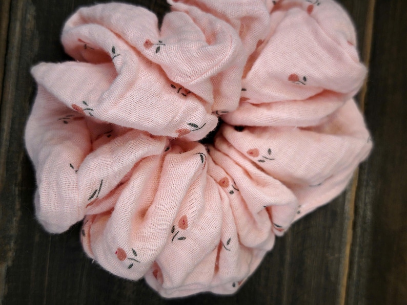 May include: A large, pale pink scrunchie with a floral design. The scrunchie is made of soft material and has small, dark green leaves and tiny red flowers. The scrunchie is designed to hold hair in a ponytail or bun.