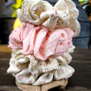 May include: A collection of fabric scrunchies in a variety of colours and patterns. The scrunchies feature floral prints in pink and cream, solid pink, and a tan polka dot design. The scrunchies are stacked on a dark wooden surface.