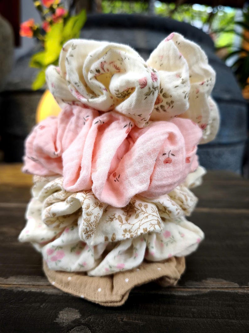 May include: A collection of fabric scrunchies in a variety of colours and patterns. The scrunchies feature floral prints in pink and cream, solid pink, and a tan polka dot design. The scrunchies are stacked on a dark wooden surface.