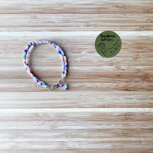 May include: A beaded bracelet with a variety of colorful beads in shades of pink, blue, yellow, and green. The bracelet has a silver clasp and a small charm. The background is a light brown wooden surface. A green sticker with the text "Handmade with love" is also visible.
