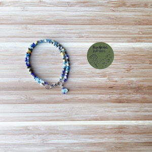 May include: A wrap bracelet made of blue, yellow, and white beads. It features a silver-coloured clasp and a small charm. A green sticker with the words "Handmade with love" and a rose is visible.
