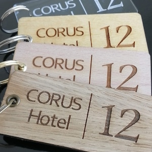 Pode incluir: Quatro chaveiros de madeira com o texto "Corus Hotel" e o número "12" gravado neles. Os chaveiros estão empilhados uns sobre os outros, com o chaveiro superior sendo um chaveiro de plástico transparente.