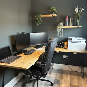 May include: A corner office workspace with a wooden desk and black hairpin legs. The desk has two computer monitors, a laptop, a printer, and a black office chair. The walls are painted a dark grey and there are two wooden shelves with plants on them.