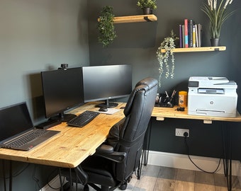 May include: A corner office workspace with a wooden desk and black hairpin legs. The desk has two computer monitors, a laptop, a printer, and a black office chair. The walls are painted a dark gray and there are two wooden shelves with plants on them.
