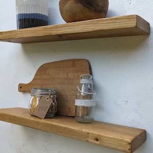 May include: Two wooden shelves mounted on a white wall. The top shelf has a clear glass vase and a wooden bowl. The bottom shelf has a wooden chopping board, a glass jar with dried fruit, and a clear glass bottle with a white string tied around it.