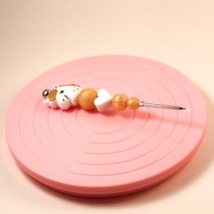 Peut inclure: Un charme de licorne blanc avec des accents dorés est enfilé sur un fil avec des perles dorées et blanches. Le charme et les perles sont sur une table tournante rose.