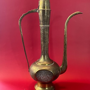 May include: A vintage brass water pitcher with intricate floral and geometric engravings. The pitcher features a long, curved spout, a handle, and a decorative lid. The base is round and sits on a small pedestal.
