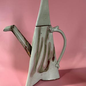 Könnte beinhalten: Eine einzigartige, geometrische Keramik-Teekanne mit einem hohen, dreieckigen Körper und einem spitzen Deckel. Die Teekanne hat eine weiße Glasur mit Streifen in Rosa, Braun und Grün. Sie hat einen gebogenen Griff und einen Ausguss.