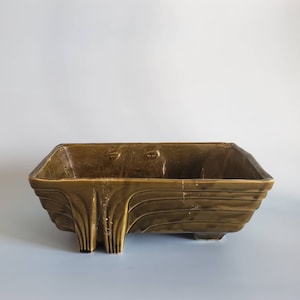 May include: A brown ceramic planter with a rectangular shape and a wavy design. The planter has four legs and a textured surface.