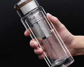May include: A double-walled glass tea infuser bottle with a stainless steel lid featuring a wood grain accent. The bottle includes a removable fine-mesh stainless steel filter basket inside, designed for steeping loose leaf tea or other beverages.