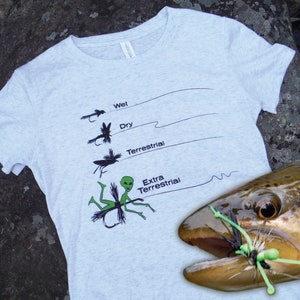 May include: A grey t-shirt with a graphic of different types of fishing flies, including a green alien fly labeled "Extra Terrestrial". A brown trout fish is biting on a green fly.